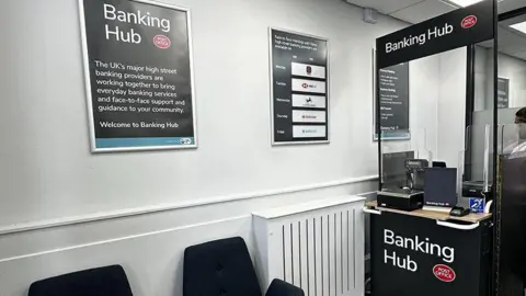 The inside of a Banking Hub. On the right is a counter with signage at the bottom and top saying "Banking Hub". There are black chairs on the left near to it.