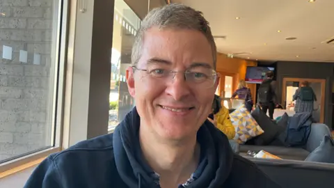 Richard has short grey hair and glasses and wears a blue hoodie.