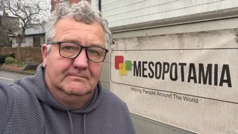 Richard Newbold, in a grey hoodie, looking at the camera, stood next to a van with a Mesopotamia banner on it