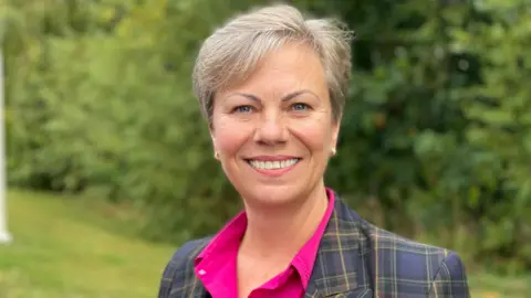 A woman with cropped-grey hair, a pink blouse and a tartan blazer, standing on a lawn in front of a row of tall bushes