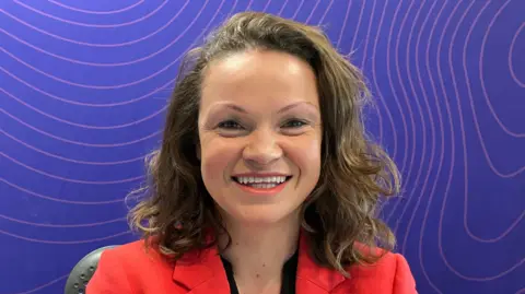 BBC A woman with brown hair. She is standing smiling at the camera. She is stood in front of a purple wall