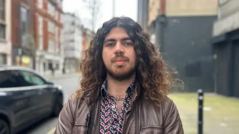 Theo has shoulder length curly hair, he wears a brown leather jacket, patterned paisley print shirt, and is looking straight at the camera, and is standing along a street with period buildings on the left, and a paved area on the right.