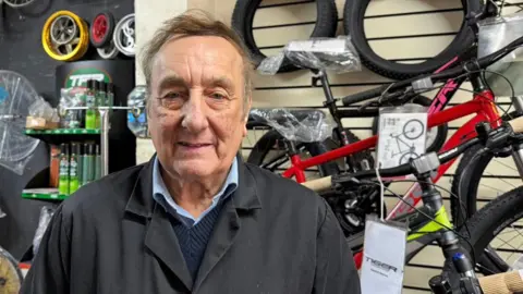BBC/Grace McGrory A man smiling at the camera as he stands in front of a rack of bikes. He is wearing a dark-grey overall-style jacket, a navy jumper and a blue shirt, which has a blue collar poking out. Behind him, to the right hand side, are three bicycles stacked up and against the wall. They have red, grey, black and pink frames. To the left are spray cans and lots of different-sized wheels on the wall. 