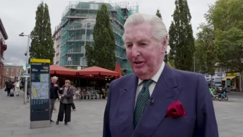 A man wears a white shirt, green spotted tie, navy suit jacket and red pocket square. He has white-blonde hair and is standing in a pedestrianised area. Behind him two people look at a map. 