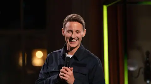 Phil McIntyre Television A man stands, smiling. He is wearing a navy jacket and is holding a microphone. He has short brown hair.