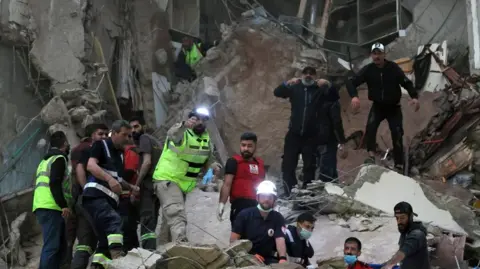 A number of rescue workers, some with helmets and head torches, others with face masks, work in the rubble of a destroyed building