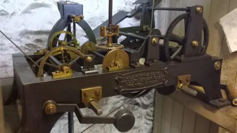 Supplied The clock cogs and gears mounted inside a metal casing with a plaque on the front