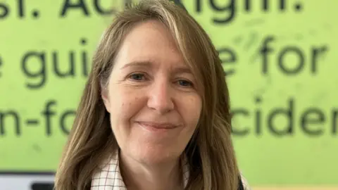 A close-up portrait shows a person standing in front of a bright green background with blurred writing that appears to reference farm safety or guidance. The person has shoulder-length hair and is wearing a collared shirt under a darker outer layer, with the focus clearly on the face while the background text remains out of focus.