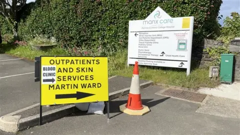 A temproary yellow sign saying 'outpatients and skin services, blood clinic, Martin Ward' with a big arrow pointing right. In the background is a permanent sign for Ramsey Cottage Hospital , signposting the various wards and units.