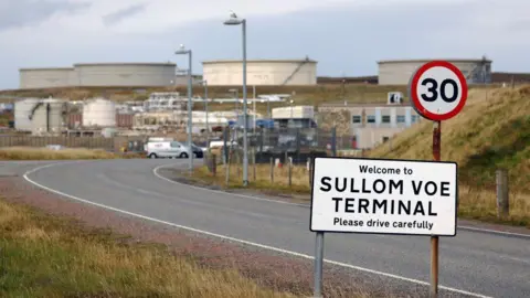 A 30mph sign which says 'Welcome to SULLOM VOE TERMINAL - Please drive carefully', with oil and gas infrastructure in the background after a winding road.