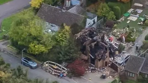 Drone photo shows destroyed buildings with emergency vehicles parked outside. Nearby homes appear undamaged