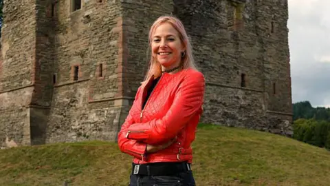 BBC/Rare TV Prof Alice Roberts wearing a red leather jacket standing in front of a castle