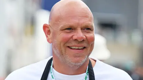 Tom Kerridge in a white t-shirt and wearing a black chef's apron. There are people walking in the background. He is standing in a Formula One paddock.