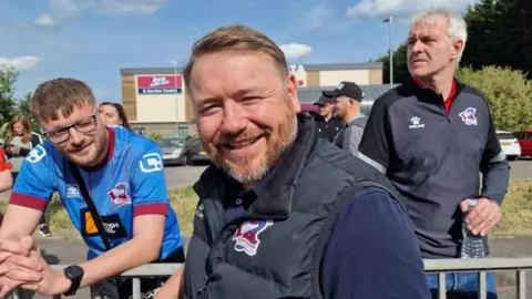 A man in a blue polo shirt with a black Scunthorpe United body warmer is smiling at the camera in front of a railing. He has short brown hair and a short brown beard and moustache. You can see a B&M behind him and several other people in Scunthorpe United tops behind the railing.