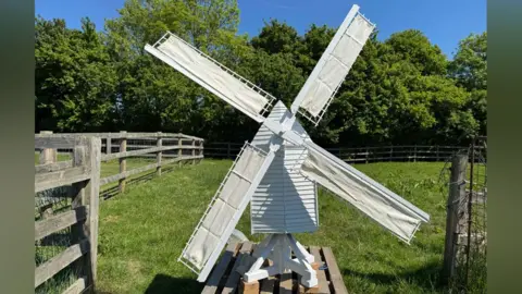 Cambridge Past, Present and Future A white replica of Bourn Windmill. It has white sails in an X-shape attached to its peaked roof. It has a white slatted body and is resting on a white X-shaped base on a pallet. Behind the model is grass and trees.