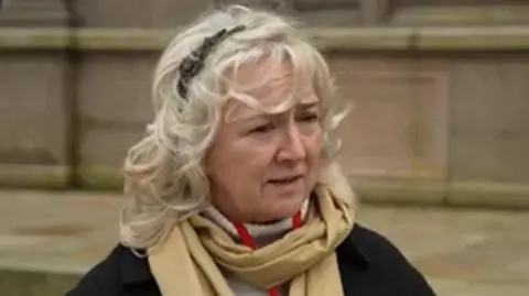 Deputy Karen Wilson is wearing a black jacket and a beige scarf. She has blonde hair and is stood outside a stone building