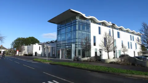 Trevor Bevins A modern white office building with a distinctive wave-shaped roof and a glass-fronted entrance stands beside a road under a clear blue sky. Trees line the pavement outside the three-storey building.