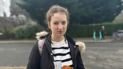 19-year-old student Florence Snoxell stood on the street with a road behind her. She is wearing a black and white stripped jumper and black coat and is hold a DNA kit and a packet of chocolate. 