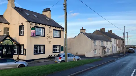 The Stag Inn in Crosby. The traditional-looking two-storey building is by the main road. It is painted cream with black detailing and sash windows. 