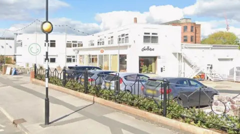 A screenshot from Google Maps of a white building with a car park outside. There is an outdoor seating area to the left of the building.