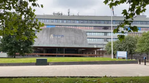 A six-storey office building with bands of glass windows. protruding out of the building in a raised Portland stone ovoid window-less council chamber. A lawn and path can be seen in the foreground.