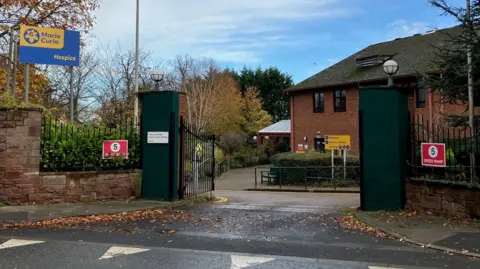 Marie Curie hospice is a two-storey brick building set behind a sandstone wall and large gates. A blue sign and yellow sign has the writing Marie Curie Hospice. There are two red traffic warning signs which say 5 speed ramp.
