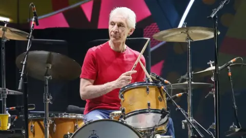 Reuters A colour photo shows a musician sitting at a drum kit on stage, wearing a red T-shirt and holding drumsticks, surrounded by cymbals, microphones and a bass drum against a bright, patterned backdrop.