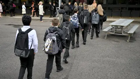 Getty Images A line of school children carrying bags walking across a playground. 