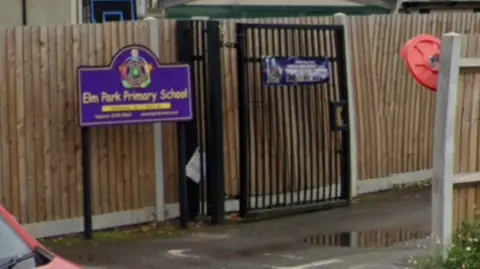 Google StreetView image showing a row of wood fencing and black gates at the entrance to the school. In front of it, a purple and yellow sign bears the Elm Park Primary School name, logo and contact information
