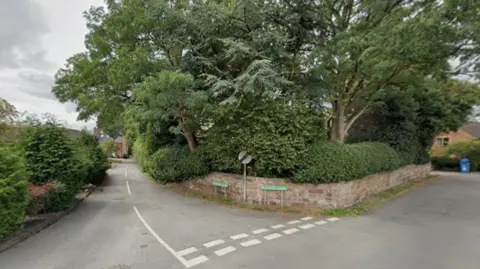 Google A corner of two residential streets, with green road signs and a white and black traffic sign.