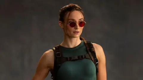A photo shows Sophie Turner as Lara Croft. She wears red-tinted sunglasses, her hair tied back. She wears a green vest top and tight, thigh-length shorts. One of her fingerless-gloved hands rests on a pistol in a thigh holster.