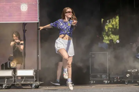 Getty Images A woman in a tartan transmt football top dances on a stage weraing a short white skirt and sunglasses.