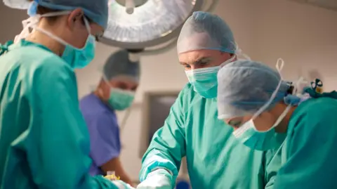 Getty Images Surgeons operating in theatre in green surgical gowns and face masks and head coverings with large lamp overhead