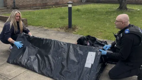 A man and a woman are crouched down on the floor at either end of the Sleep pod - a long, black, single-person tent in the shape of a triangle.
There is a black dog laying down behind the pod
The woman has long blonde hair, wears a dark blue hoodie and blue latex gloves. The man wears a police uniform and latex gloves. Both hold opposite ends of the triangular shelter.