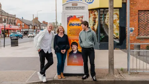 KCOM Three people - a man and his parents - stand either side of a cream mid-20th Century telephone box in a broad high street. The phone box is decorated with a colourful poster featuring a picture of the actor and the words: "Hometown heroes - acclaimed actor Rob Aramayo has blazed a trail from right here in East Hull to Hollywood". The man is wearing a green hoodie, black jeans and black trainers. He is smiling as he looks at his parents. His father is leaning against the phone box and wearing a cream jacket, white shirt, black jeans and white trainers. His mother, who has strawberry blonde hair, is wearing a blue top, matching jeans and brown boots.