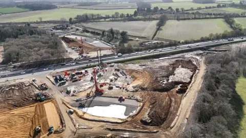 Shaun Whitmore/BBC The construction site with cranes, lorries and sandy excavations, next to the road with cars travelling along it.
