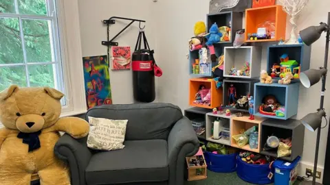 A room with a large brown teddy bear next to a grey armchair near a window. On the right there are boxes of different colours including toys.