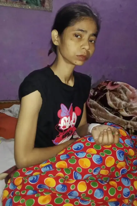 Shaheen Malik An extremely thin and gaunt-looking woman sits on a bed with a mattress pulled over her legs.