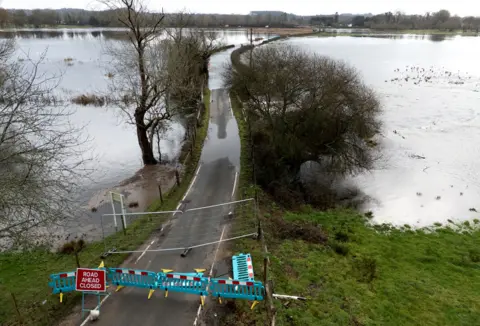 PA Media A 'road closed ahead' sign and green barriers block a road flooded with high waters on either side of it near to Harbridge in Hampshire, taken on Friday.