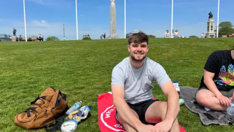 Jude Harris sat on a red towel, on the grass, smiling at the camera. Beside him is a brown rucksack.