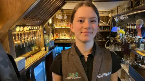 BBC/Seb Cheer A young woman, Hope, smiles at the camera. She is wearing a brown waistcoat. She stands in a bar area.