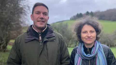 A man and a woman smiling at the camera. The man is wearing a khaki waterproof coat and fleece. The woman has shoulder length brown hair and is wearing a leather-look jacket and blue and purple scarf. In the background can be seen green fields, hedges and a grassy hill.