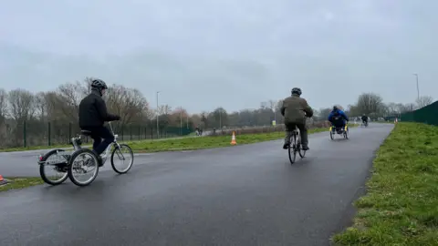 4 people are seen riding into the distance on trikes and bikes. The cycle track is a wide tarmac surface with grass either side and cones lining it.