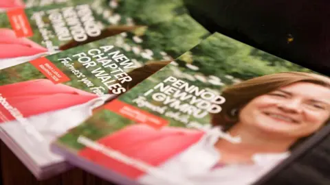 A set of manifestos laid out of a surface with Eluned Morgan's face on the booklets. The manifestos says "Pennod Newydd i Gymru" and "A New Chapter for Wales".