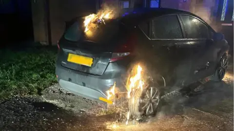 North Wales Fire and Rescue Service Flames around a rear tyre and top of a black car parked in darkness near a house in Rhosllanerchrugog