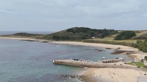 BBC Aerial view of a quiet coastal bay with clear blue-green water, sandy and rocky beaches, and a small stone pier extending into the sea. A cluster of white buildings sits among lush gardens and trees on the shoreline, while rolling green hills and wooded areas rise in the background beneath a lightly clouded sky.