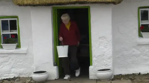 A screen grab of Margaret Gallagher when she was filmed by the BBC in 2019 leaving her cottage to fill a bucket of water from her well.  She is wearing a red cardigan over a yellow top, dark trousers and white trainers.