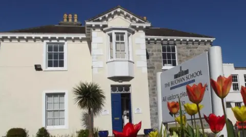 A white building that looks like a home with a blue door and a palm tree outside. A white sign in the foreground reads The Buchan School. You can also see red and yellow tulips.