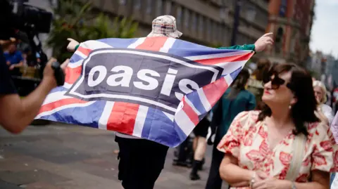 PA Media A man stands with his arms out, he holds a Union Jack flag with Oasis written across it.