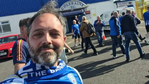A man smiling at the camera, he is wearing a blue and white scarf and top, he has dark hair and beard
